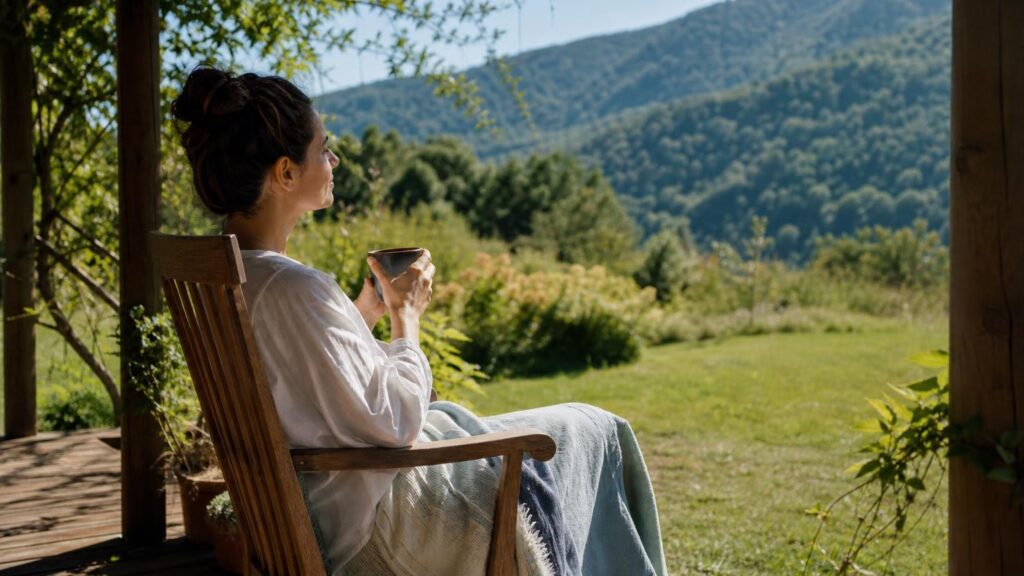 Woman in a white shirt sitting in a wooden chair on a porch, holding a mug and looking out at lush green mountains under a bright sun.
